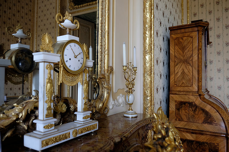 Old clock with golden elements on mantelpiece in front of a mirror with golden border, in the background a wooden writing cabinet.