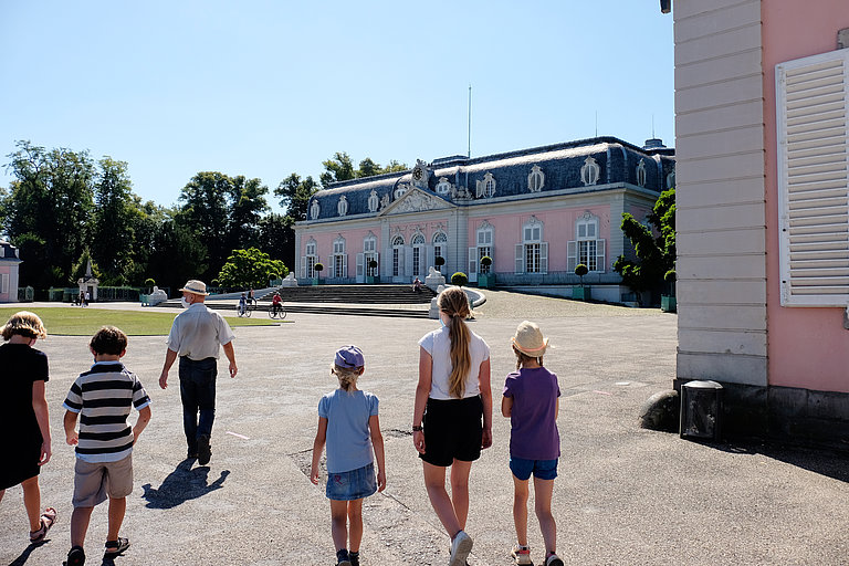 Fünf Kinder folgen einem Mann mit Hut in Richtung Corps de Logis