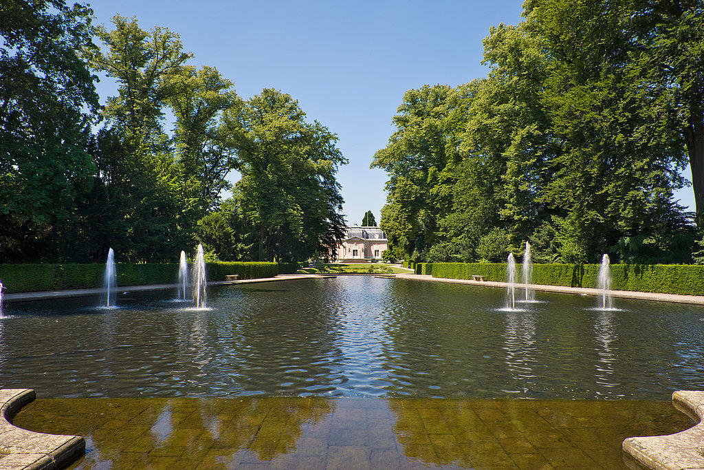 Wasserbecken mit Wasserfontänen im französischen Garten mit Blick auf das rosafarbene Corps de Logis.