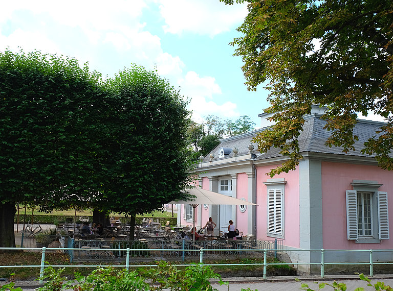 Blick auf eine bestuhlte Terasse mit Bäumen, rechts daneben das rosafarbene Gebäude des Schlosscafés