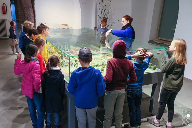 Eine Gruppe von Kindern steht um einen Schaukasten mit der Miniaturansicht von Schloss Benrath herum.