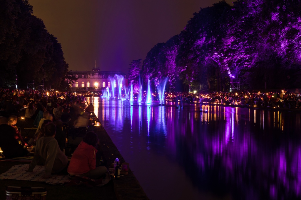 Violet beleuchtete Bäume, Menschen um den Spiegelweiher sitzend aus dem blau leuchtende Wasserfontönen entspringen